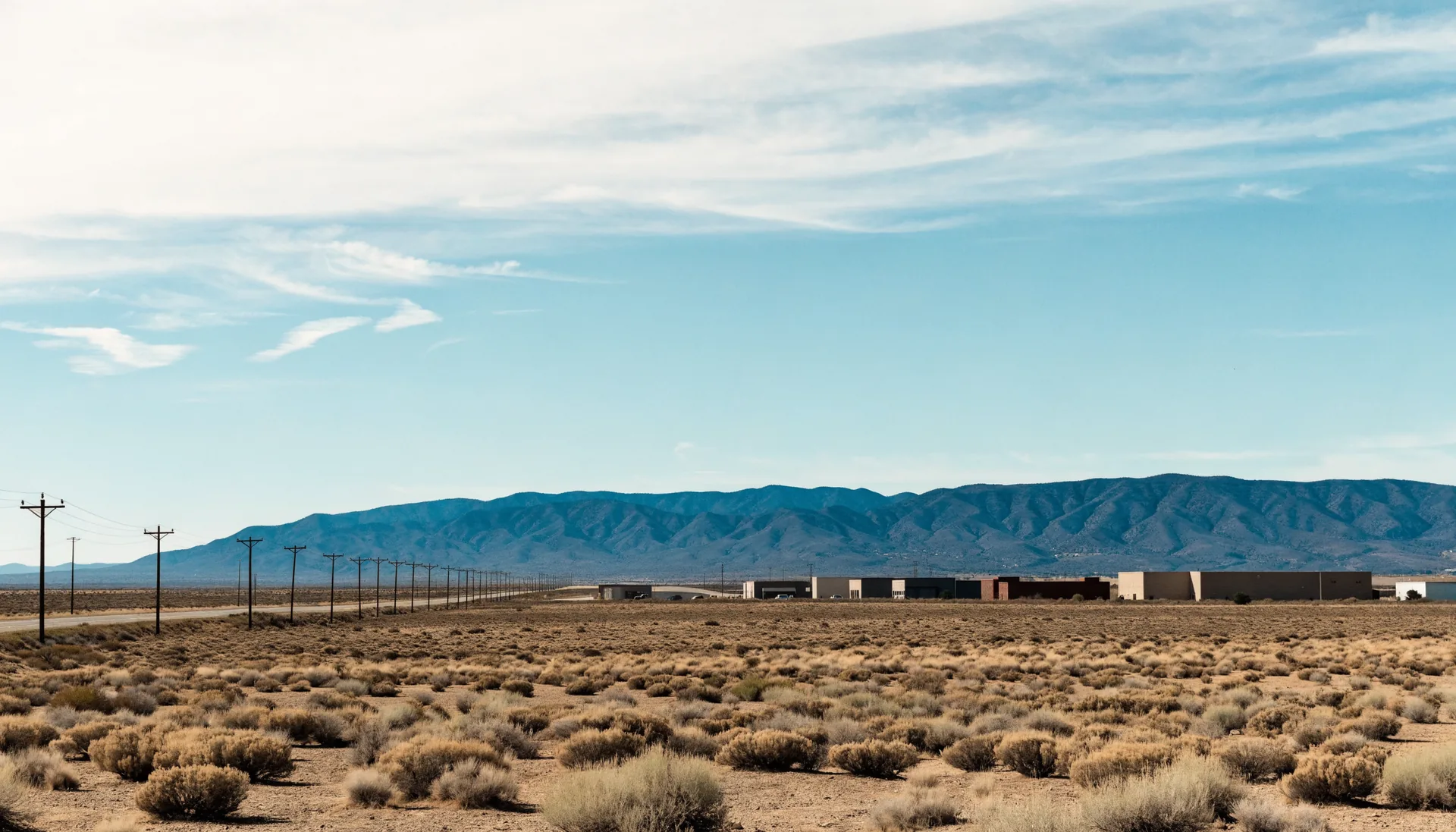 High desert scrub on the edge of a southwestern city with an overhead cable line running toward low industrial buildings and a long mountain ridge behind them