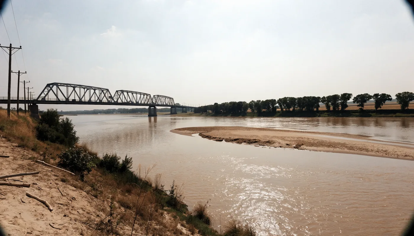 A steel truss bridge carrying a highway over a wide muddy midwestern river with farmland beyond