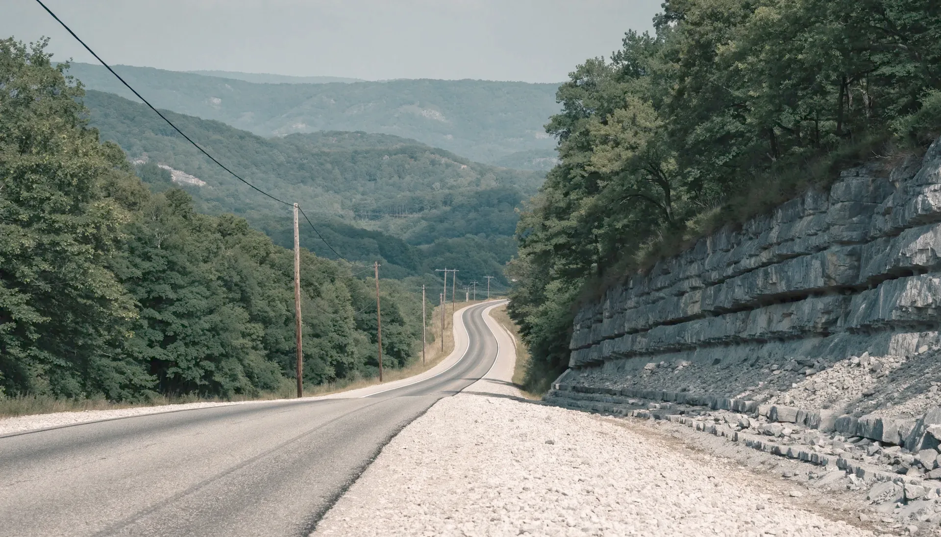 A two lane highway cutting through wooded Ozark hills past a raw limestone rock cut with an overhead cable line following the road